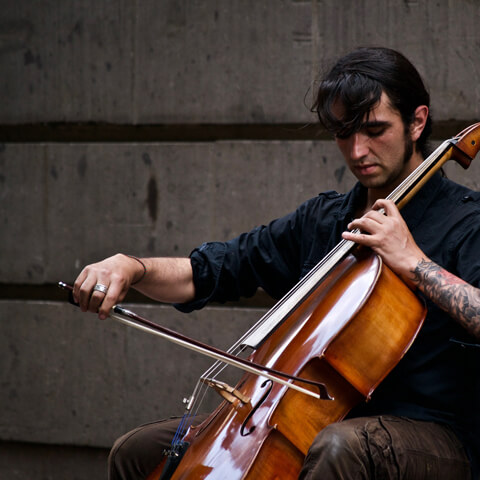 A tattoed man plays cello in front of a concrete wall. Photo by Jannel Fosati (Unsplash)